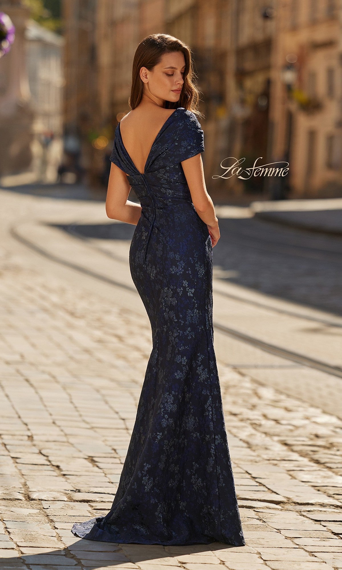 A woman stands on a cobblestone street wearing the La Femme 33571 long formal dress, a navy blue, floor-length, off-the-shoulder gown with a subtle floral pattern.